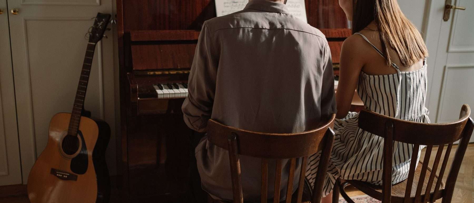Eine Akustikgitarre lehnt ungenutzt an der Wand, während zwei Personen Klavier spielen